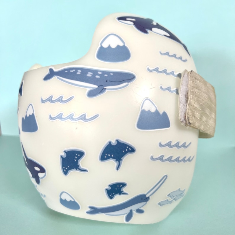Pu&ograve; includere: Casco bianco con un design a tema oceanico blu e grigio. Il casco ha un cinturino laterale e presenta balene, narvali, razze, montagne e onde.