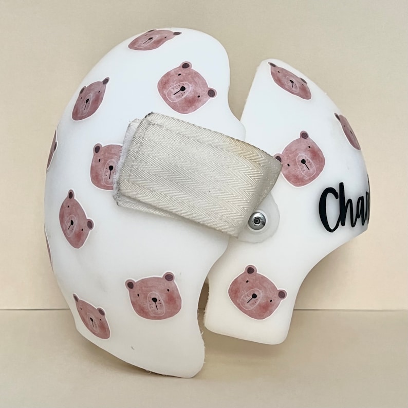 May include: White helmet with a brown bear pattern and the name "Charlie" written in black on the side. The helmet has a white strap with a velcro closure.