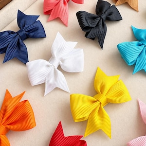 May include: A collection of hair bows made from ribbed grosgrain ribbon in various solid colours including navy blue, white, black, bright yellow, orange, pink, turquoise, and purple, each featuring a classic tied bow shape with folded loops.