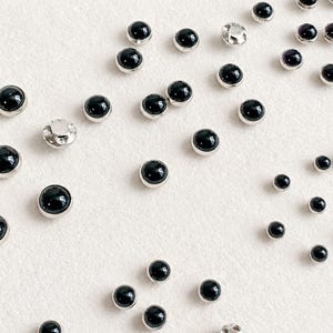Può includere: Una collezione di piccoli bottoni o borchie rotondi in stile cabochon, ognuno caratterizzato da una superficie nera liscia e lucida incastonata in un sottile bordo metallico color argento. Gli articoli sono sparsi nel riquadro in varie dimensioni.