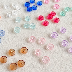Puede incluir: Una colección de botones pequeños, redondos y de dos agujeros en una variedad de colores translúcidos que incluyen naranja, transparente, azul, rosa, rojo, verde, amarillo, morado y marrón, dispersos en pequeños grupos de dos o tres piezas.