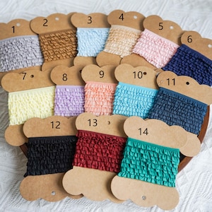 May include: A collection of 14 spools of ruffled trim in various colors, including light blue, pink, yellow, and brown. Each spool is labeled with a number from 1 to 14.