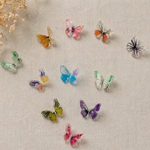 May include: A collection of colorful, translucent butterfly embellishments in various sizes and hues, including orange, pink, green, blue, and purple. The butterflies are scattered on a textured, off-white surface, with a sprig of dried flowers in the upper left corner.