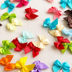 Pode incluir: Uma coleção de pequenos laços decorativos de fita de cetim em uma variedade de cores vibrantes, incluindo vermelho, verde brilhante, amarelo, laranja, roxo, marrom, azul claro e creme, espalhados sobre uma superfície lisa e clara.