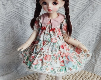 May include: A doll wearing a white dress with pink and floral accents, a pink collar, and a white lace trim. The doll has brown hair in braids and is wearing white socks and brown shoes.