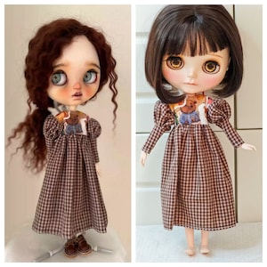 Puede incluir: Dos muñecas con vestidos de cuadros marrones y blancos con mangas abullonadas. Los vestidos tienen un estampado de oso marrón en el corpiño. Una muñeca tiene el pelo largo y rizado marrón y la otra tiene el pelo corto y liso marrón.