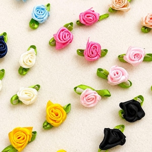 May include: A collection of small fabric ribbon roses in various colours including red, blue, yellow, pink, white, and black. Each rose features a tightly rolled centre and two small green ribbon leaves attached at the base for decoration.