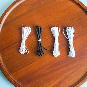 Peut inclure: Quatre paquets de cordon élastique de différentes couleurs : rose clair, noir, blanc et bleu clair. Les cordons sont soigneusement attachés avec de petits élastiques sur un plateau rond en bois.
