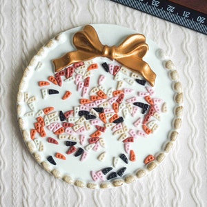 May include: A white ceramic round ornament with a gold bow on top. The ornament is decorated with small, colorful plastic pieces in shades of pink, orange, white, and black.