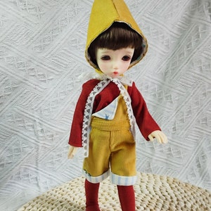 Könnte beinhalten: Eine Puppe in einem gelben Hut mit einem bordeauxfarbenen Bommel, einer roten Jacke, einer gelben Cord-Latzhose und roten Socken. Die Puppe hat einen hellen Teint und dunkles Haar. Das Outfit umfasst einen weißen Spitzenkragen und blau-weiß gestreifte Manschetten.