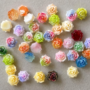 May include: A collection of small, three-dimensional rose-shaped cabochons in a variety of colors including pink, yellow, blue, green, orange, white, and deep red. Each rose features intricate petal details and a glossy, semi-translucent finish.