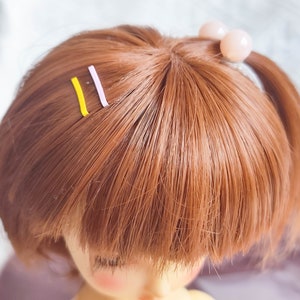 May include: A doll with long, brown hair styled in two pigtails with a pink hair tie. The hair is held in place with two bobby pins, one yellow and one purple.