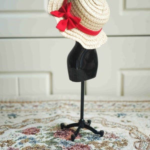 May include: A straw hat with a red bow sits on a black mannequin stand. The stand has a round base with three legs. The hat and stand are on a patterned rug.