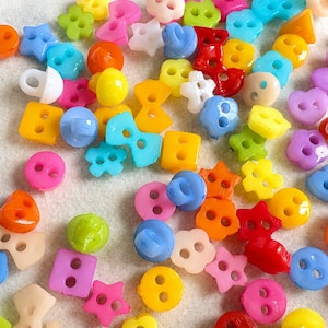 May include: A collection of small, multi-coloured plastic buttons in various shapes, including stars, flowers, squares, circles, and bows. The buttons feature two or four holes for sewing and come in a vibrant palette of red, blue, yellow, pink, orange, green, and white.
