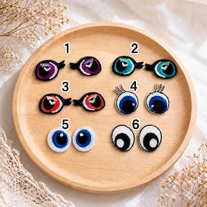 Peut inclure: Un plateau en bois présente six écussons brodés en forme d'yeux, aux designs variés. Les écussons arborent différentes couleurs, dont le violet, le turquoise, le rouge et le bleu, avec des détails noirs. Certains ont des cils, et tous sont numérotés.