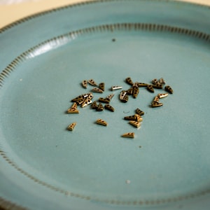 Może przedstawiać: Niebieski ceramiczny talerz z rozrzuconymi na nim małymi, złotymi metalowymi elementami. Elementy mają kształt trójkątów z małym otworem w środku.