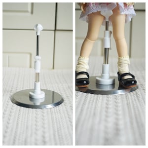 May include: White doll stand with a metal base and adjustable height. The stand is shown with a doll wearing a white dress and black shoes.