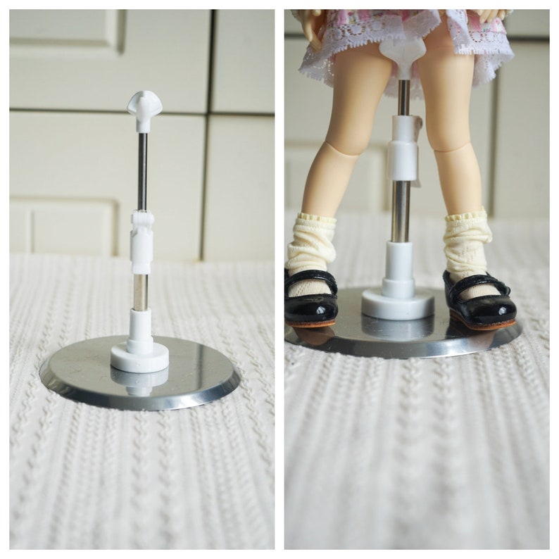 May include: White doll stand with a metal base and adjustable height. The stand is shown with a doll wearing a white dress and black shoes.