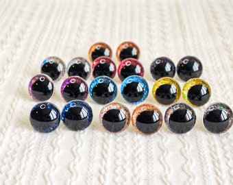May include: Assortment of colorful glitter googly eyes with black pupils. The eyes are arranged in a triangular pattern on a white surface.