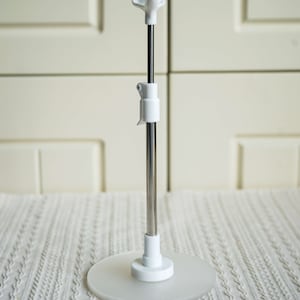 May include: A white adjustable stand with a round base and a metal rod. The stand is designed to hold a variety of items, such as phones, tablets, or books.