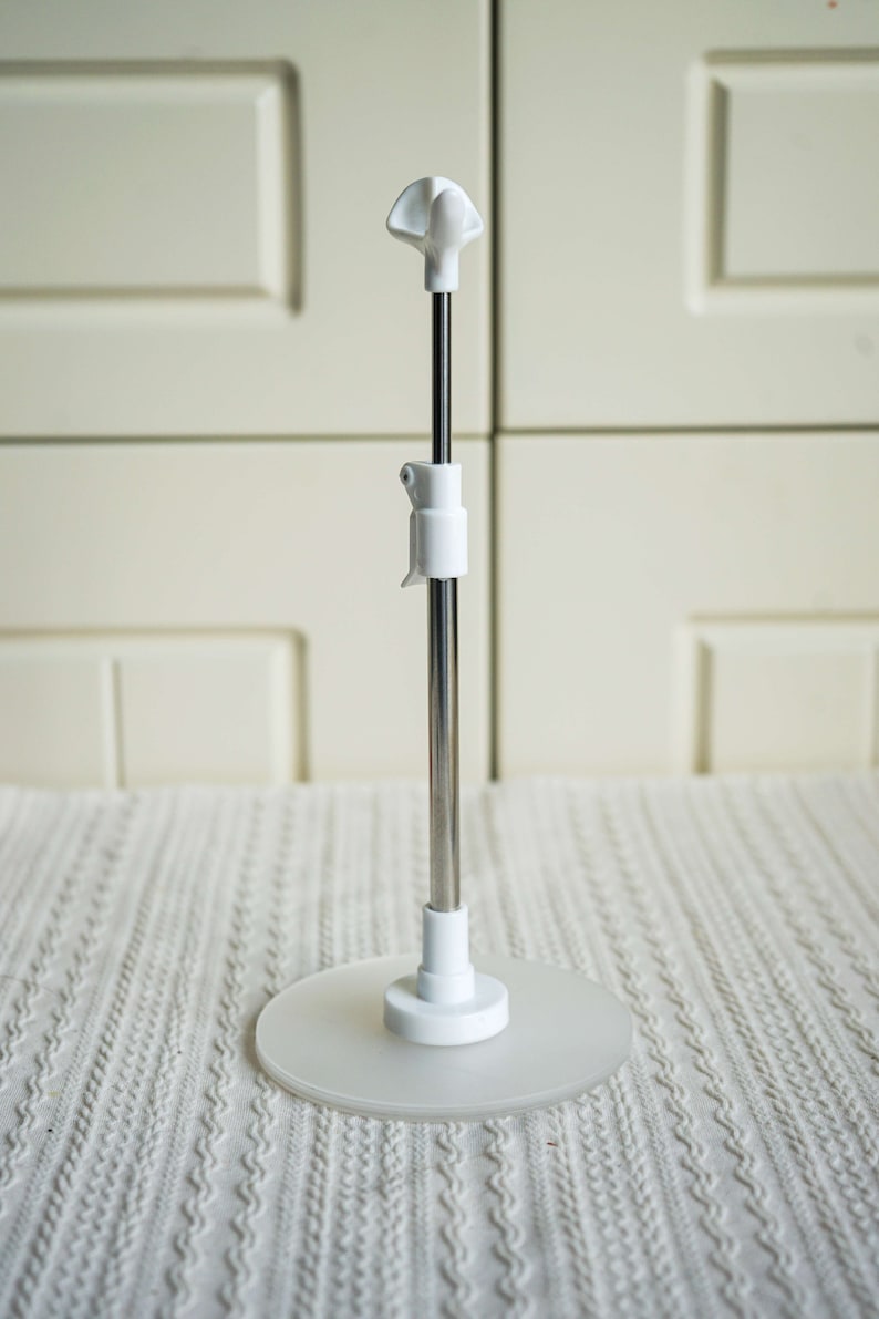 May include: A white adjustable stand with a round base and a metal rod. The stand is designed to hold a variety of items, such as phones, tablets, or books.