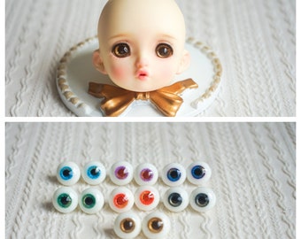 May include: A collection of 12 doll eyes in various colors, including blue, green, brown, red, and purple. The eyes are arranged in a circle on a white surface.