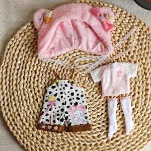 May include: A set of doll clothes featuring a pink hooded hat with a bear face, a pink and white polka dot shirt, and black and white polka dot overalls with a brown trim.