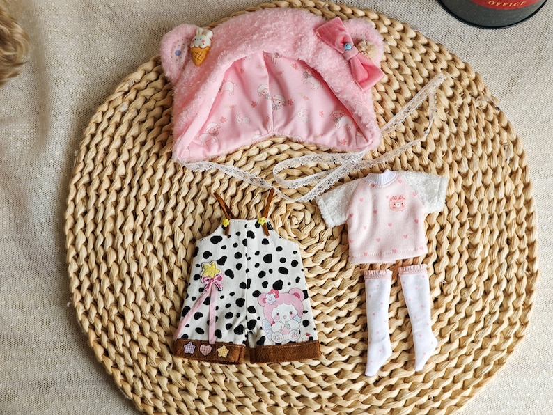 May include: A set of doll clothes featuring a pink hooded hat with a bear face, a pink and white polka dot shirt, and black and white polka dot overalls with a brown trim.
