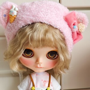 May include: A doll with blonde hair wearing a pink fuzzy hat with a bow and a small ice cream cone charm. The doll is wearing a white shirt with a pink undershirt and black and white overalls.