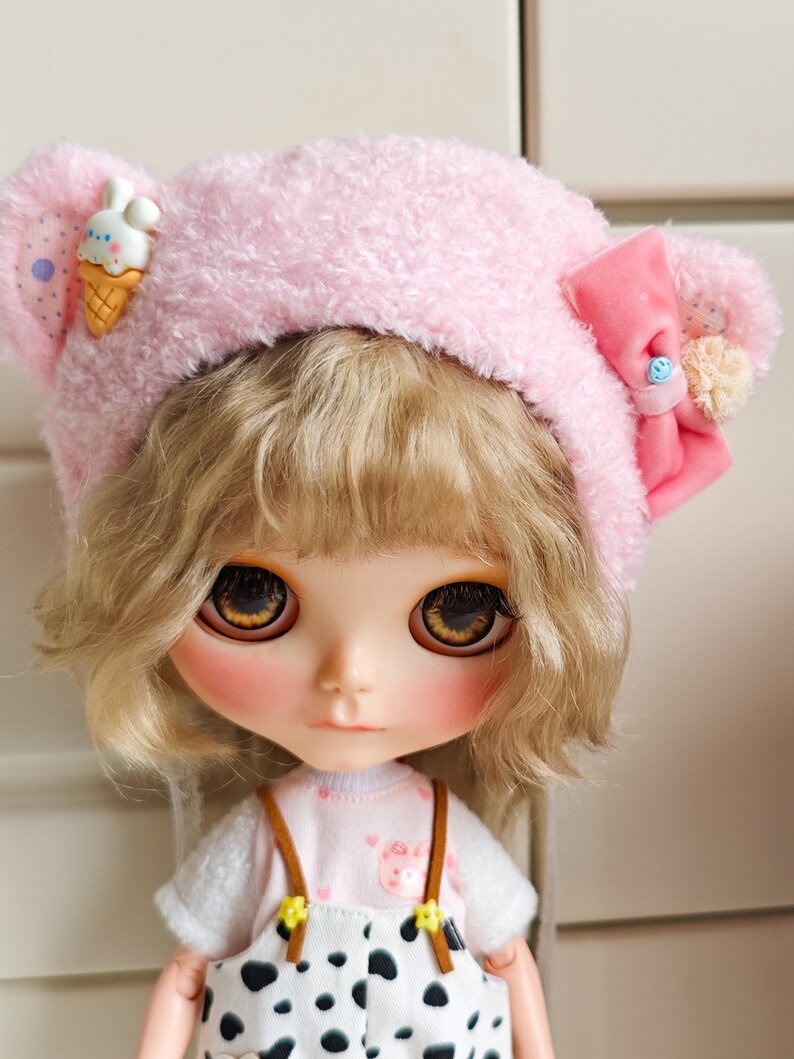 May include: A doll with blonde hair wearing a pink fuzzy hat with a bow and a small ice cream cone charm. The doll is wearing a white shirt with a pink undershirt and black and white overalls.