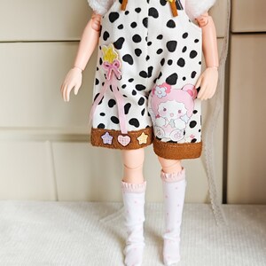 May include: A doll wearing a white and black cow print overall with brown straps and a pink bow. The overall has a brown trim and a pink and white character patch. The doll is also wearing white socks with pink polka dots.