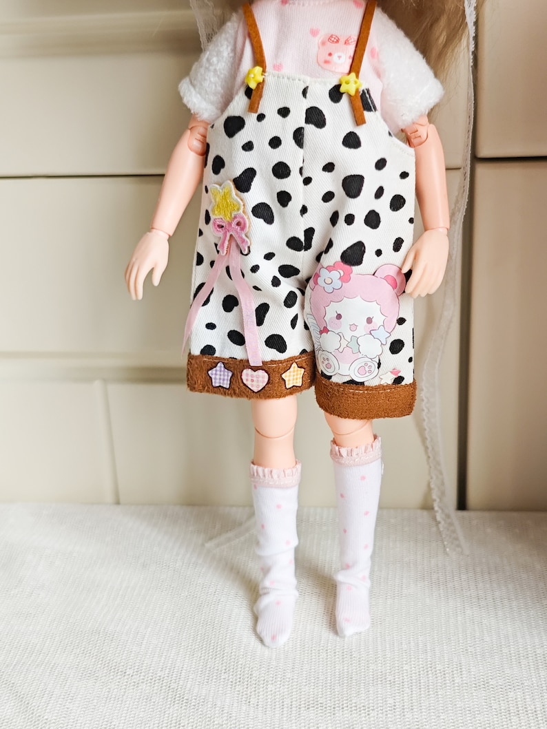 May include: A doll wearing a white and black cow print overall with brown straps and a pink bow. The overall has a brown trim and a pink and white character patch. The doll is also wearing white socks with pink polka dots.