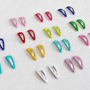Peut inclure: Une collection de vingt-quatre barrettes à cheveux en métal disposées par paires, présentant une variété de couleurs unies dont le rouge, le bleu, le vert, le jaune, le rose, le blanc et le turquoise, avec un mécanisme en métal argenté visible.