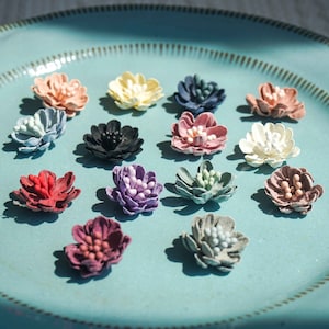 May include: Assortment of 14 small fabric flowers in various colors, including pink, purple, red, blue, green, white, and black. The flowers are arranged on a light blue plate.