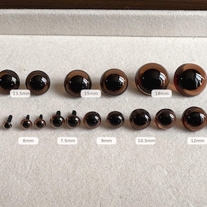 Puede incluir: Una colección de ojos de seguridad redondos y marrones para manualidades, dispuestos en dos filas. La fila superior presenta los tamaños 13,5 mm, 15 mm y 18 mm. La fila inferior muestra los tamaños 4,5 mm, 6 mm, 7,5 mm, 9 mm, 10,5 mm y 12 mm, cada uno con un pequeño poste negro.