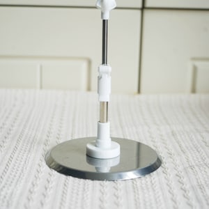 May include: A white metal display stand with a round base and a telescoping rod. The stand is adjustable in height and can be used to display a variety of items.