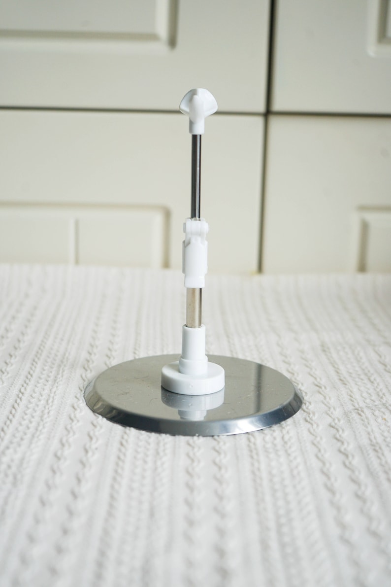 May include: A white metal display stand with a round base and a telescoping rod. The stand is adjustable in height and can be used to display a variety of items.