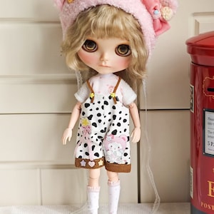 May include: A doll with blonde hair wearing a pink fuzzy hat with bear ears and a white and black polka dot dress. The doll is wearing white socks with pink trim and is standing on a white surface.