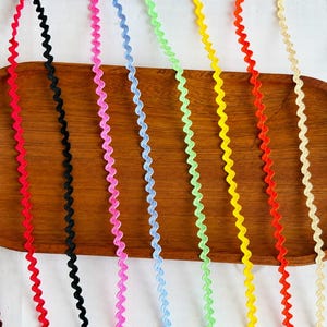 Può includere: Una collezione di dieci nastri a zig-zag colorati disposti verticalmente. I colori includono marrone scuro, rosso, nero, rosa, azzurro, verde lime, giallo, arancione, crema e turchese. Ogni nastro presenta un motivo ondulato costante.