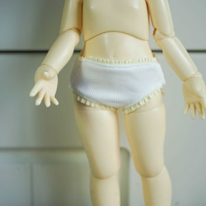 Op de afbeelding: Een witte pop die witte katoenen ondergoed draagt met een kantversiering.