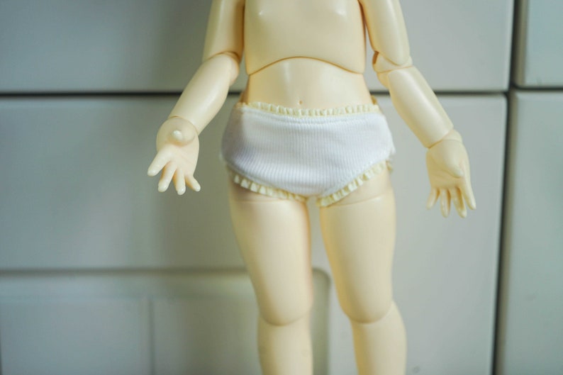 Op de afbeelding: Een witte pop die witte katoenen ondergoed draagt met een kantversiering.