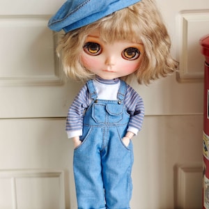 May include: A doll with blonde curly hair wearing a blue beret, a striped long-sleeved shirt, and blue denim overalls. The doll is holding a small white teddy bear.