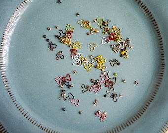 May include: A blue plate with a variety of colourful metal clasps and fasteners. The clasps are in different shapes and sizes, including heart shapes, circles, and squares. Some of the clasps are silver, gold, red, yellow, and green.