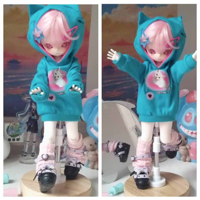 May include: A doll wearing a turquoise hoodie with cat ears and a pink and white graphic of a cat. The hoodie has the text "I love you" on the front. The doll is wearing pink and white leg warmers and black shoes with white bows.