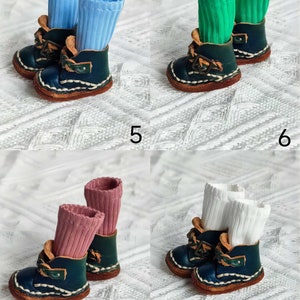 May include: Four pairs of miniature leather boots with different colored socks. The boots are dark blue with brown stitching and a buckle. The socks are light blue, green, pink, and white.