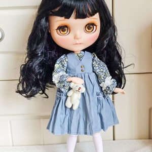 May include: A doll with long black curly hair, wearing a blue dress with a floral print and white socks. She is holding a white teddy bear.