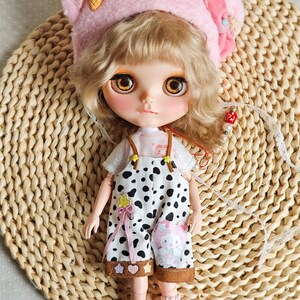 May include: A doll with blonde curly hair wearing a pink fuzzy hat with a cartoon ice cream cone on top, a white shirt with a pink and white polka dot pattern, and black and white spotted overalls with a brown trim. The doll is wearing white socks with pink polka dots and is standing on a woven straw mat.