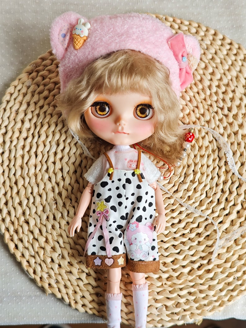 May include: A doll with blonde curly hair wearing a pink fuzzy hat with a cartoon ice cream cone on top, a white shirt with a pink and white polka dot pattern, and black and white spotted overalls with a brown trim. The doll is wearing white socks with pink polka dots and is standing on a woven straw mat.