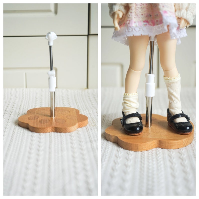 May include: A doll stand with a wooden base shaped like a paw print. The stand has a metal pole with a white plastic connector. A doll is standing on the stand, wearing a white dress, white socks, and black shoes.