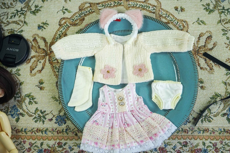 Op de afbeelding: Een set poppenkleding inclusief een wit gebreid vest met roze bloemaccenten, een roze en witte jurk met kantversiering, witte sokken en witte ondergoed met gele versiering.